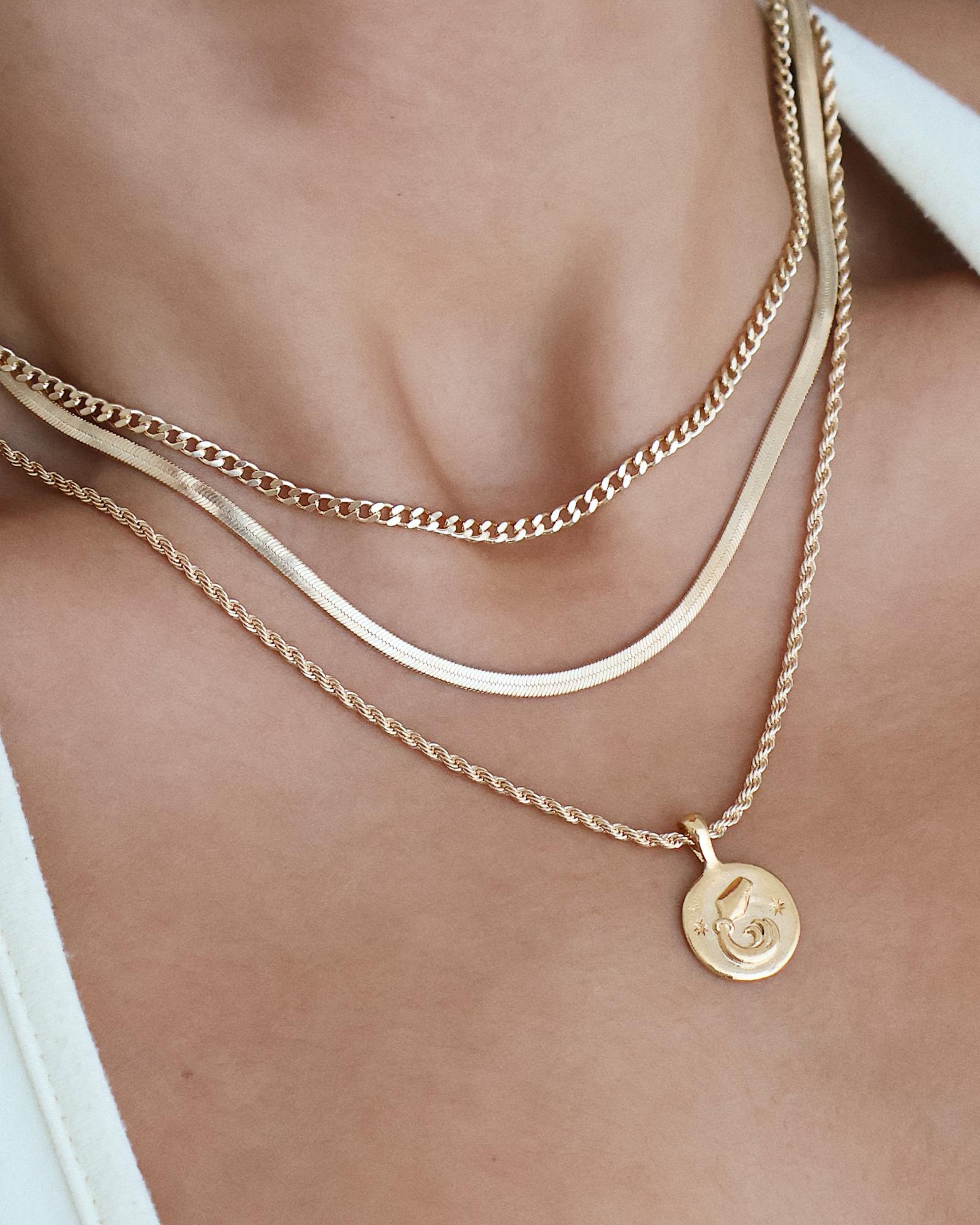Close-up detail image of layered gold necklaces worn on the neckline, featuring a flat gold snake chain, a medium gold curb chain, and a fine gold chain with a round coin pendant. The image highlights how different gold chain styles sit together at staggered lengths, creating a balanced layered look. Designed for women seeking versatile gold necklaces and inspiration for effortless layering, helping solve the challenge of mixing chain styles without overwhelming the outfit.