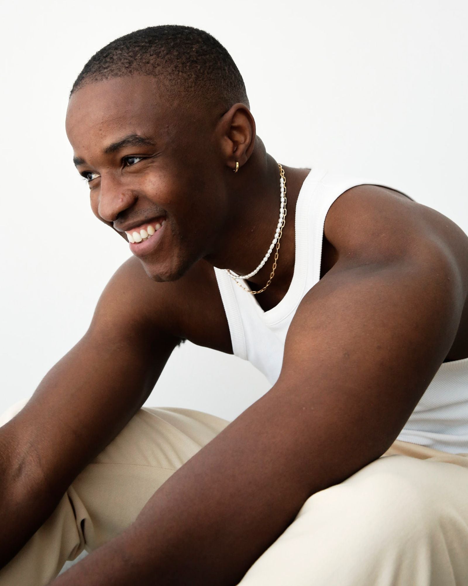 Lifestyle image of a male model wearing a short freshwater pearl necklace layered with a fine gold chain and a small gold hoop earring, styled with a white tank top. Designed for men seeking a modern pearl necklace that offers a relaxed, gender-neutral approach to jewellery, ideal for everyday wear, layering, and adding understated elegance to casual outfits.