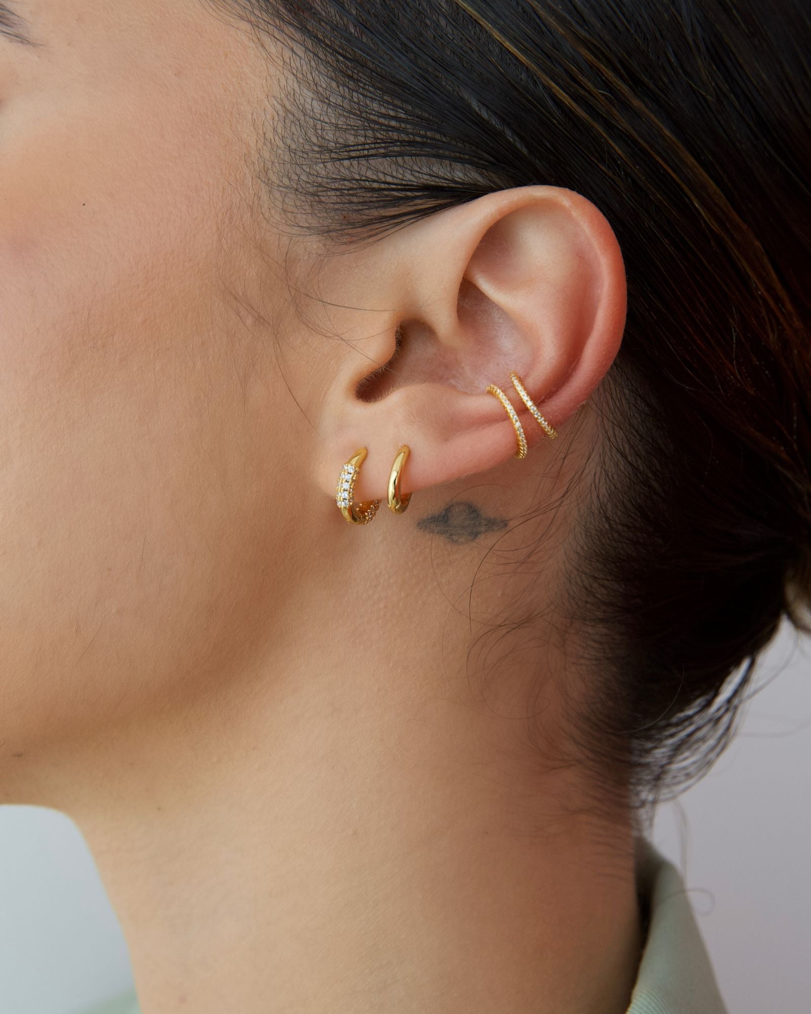 Side profile close-up of a woman’s ear styled with a layered gold ear stack, featuring two slim no-piercing ear cuffs on the upper ear and two small gold hoop earrings in the lobe, one accented with clear gemstones. The image highlights minimalist gold ear jewellery designed for people who want a curated, stacked ear look without additional piercings, offering an easy way to add subtle sparkle, balance, and everyday elegance to an ear stack.