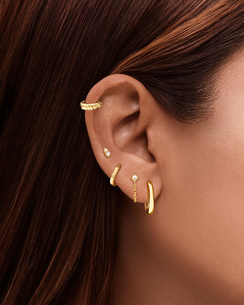 Close-up of a woman’s ear styled with the Xanthe textured gold huggie hoop in the lobe, paired with minimalist gold studs and an ear cuff. The image shows how textured gold huggie earrings sit close to the ear for a modern, layered ear stack. Designed for women seeking everyday gold hoops that add subtle statement and dimension while remaining lightweight, comfortable, and suitable for sensitive ears.