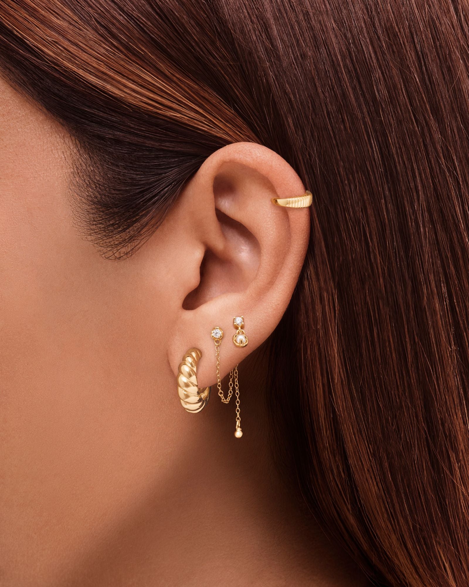 Close-up of a woman’s ear styled with layered gold earrings, featuring twisted gold hoop earrings, delicate gold chain drop studs, and a minimalist gold ear cuff. Designed for women seeking everyday gold earrings and modern ear stacking inspiration, this image shows how small gold hoops and dainty drop earrings can be layered to create a polished, versatile look for multiple piercings and effortless daily wear.
