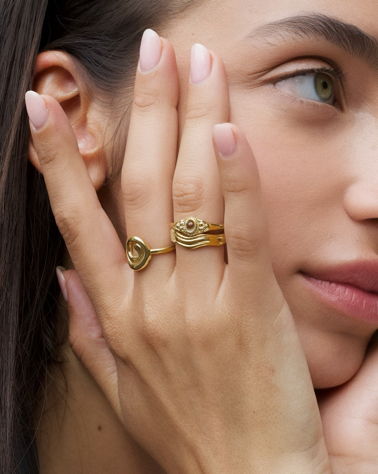 Product image showing a close-up of a woman wearing layered gold rings on her fingers, including a spiral gold ring, a wave-style stacking ring, and an oval stone gold ring, with a natural manicure and minimal makeup. Designed for women seeking everyday gold rings, gold stacking rings, and versatile jewellery styling, this image demonstrates how minimalist gold rings can be worn together to create an elevated, cohesive look that transitions easily from casual to refined wear.