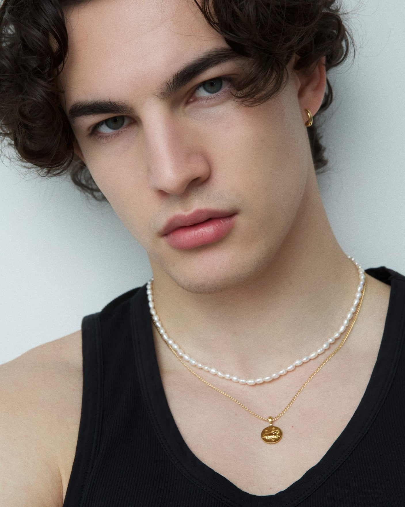 Close-up portrait of a model wearing a short freshwater pearl necklace layered with a fine gold chain and round gold pendant, paired with a small gold hoop earring. Designed for women and men seeking a modern pearl necklace with a refined, gender-neutral aesthetic, ideal for everyday wear, layering, and elevating minimal outfits with timeless elegance.