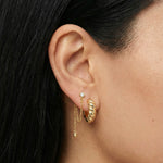 Close-up product photo of a woman’s ear with warm medium skin tone and sleek dark hair, styled with layered gold earrings. The ear features a twisted gold huggie hoop in the lobe and a delicate gold chain drop earring anchored by a small clear stone stud, finished with a tiny gold bead at the end of the chain. This image is for customers looking for refined gold earrings that combine subtle texture with soft movement for everyday wear. It shows how minimalist gold huggies and chain earrings can be paired to