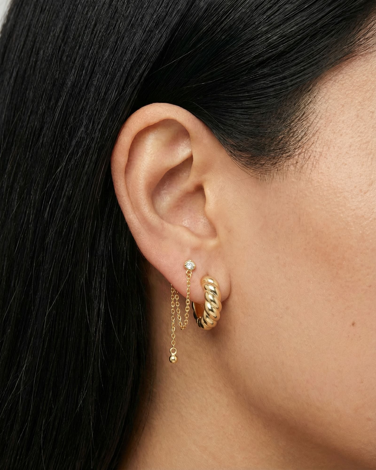 Close-up product photo of a woman’s ear with warm medium skin tone and sleek dark hair, styled with layered gold earrings. The ear features a twisted gold huggie hoop in the lobe and a delicate gold chain drop earring anchored by a small clear stone stud, finished with a tiny gold bead at the end of the chain. This image is for customers looking for refined gold earrings that combine subtle texture with soft movement for everyday wear. It shows how minimalist gold huggies and chain earrings can be paired to