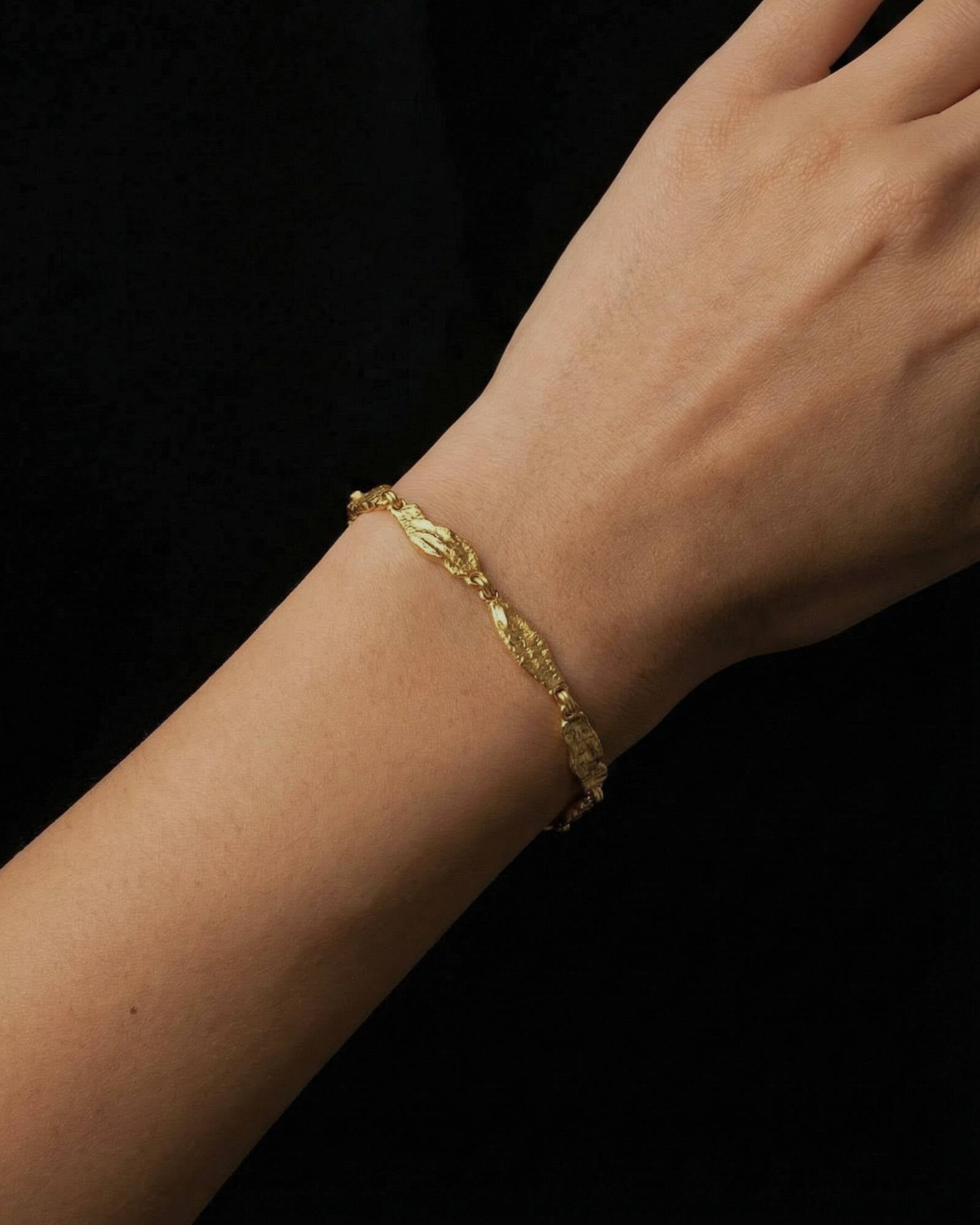 Close-up of a woman’s wrist wearing a slim gold bracelet with textured, organically shaped links, photographed against a dark background. The image clearly shows the bracelet’s warm gold tone, detailed surface finish, and how it sits naturally on the wrist. Designed for women seeking refined, everyday gold bracelets that add subtle interest without overpowering an outfit, this image helps customers understand scale, fit, and how a delicate gold bracelet elevates a minimal, modern look.
