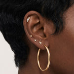 Close-up product photo of a woman’s ear with deep brown skin and natural textured hair, styled with layered gold earrings across multiple piercings. The ear features a large smooth gold hoop in the lobe, several small gold studs with clear stones in the lower and mid ear, including a tiny round stud, a cross-shaped stud, and a pavé-style stud, as well as a delicate cluster of small clear stones in the upper cartilage. This image is for customers looking for versatile gold earrings that suit multiple piercin