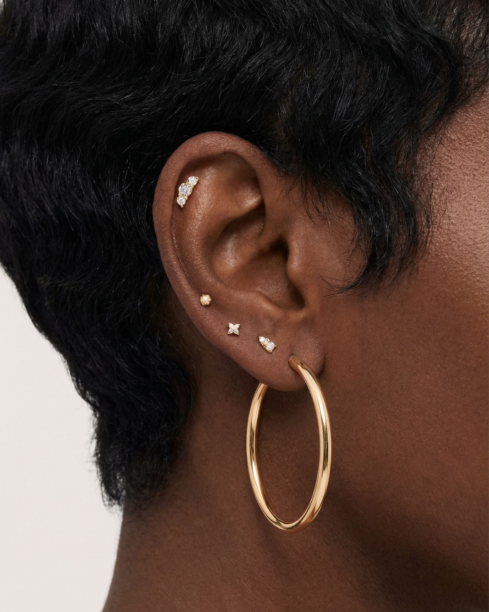 Close-up product photo of a woman’s ear with deep brown skin and natural textured hair, styled with layered gold earrings across multiple piercings. The ear features a large smooth gold hoop in the lobe, several small gold studs with clear stones in the lower and mid ear, including a tiny round stud, a cross-shaped stud, and a pavé-style stud, as well as a delicate cluster of small clear stones in the upper cartilage. This image is for customers looking for versatile gold earrings that suit multiple piercin