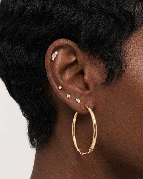 Close-up product photo of a woman’s ear with deep brown skin and natural textured hair, styled with layered gold earrings across multiple piercings. The ear features a large smooth gold hoop in the lobe, several small gold studs with clear stones in the lower and mid ear, including a tiny round stud, a cross-shaped stud, and a pavé-style stud, as well as a delicate cluster of small clear stones in the upper cartilage. This image is for customers looking for versatile gold earrings that suit multiple piercin