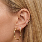 Close-up product photo of a woman’s ear with fair skin and straight blonde hair, styled with layered gold earrings across multiple piercings. The ear features a large smooth gold hoop in the lobe, a pavé gold huggie set with clear stones in a second piercing, a small gold stud with a clear stone in the mid ear, and a slim pavé gold huggie in the upper cartilage. This image is for customers looking for statement gold earrings that still feel refined and wearable for everyday use. It shows how classic gold ho
