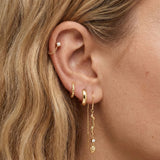 Close-up side profile of a blonde woman’s ear wearing layered gold jewellery, featuring a gold huggie hoop, a fine chain threader earring with a sculptural wavy bar and small crystal drop, and a delicate cartilage stud. Designed for women who like modern, minimal ear stacks, this image helps customers visualise how the gold drop earrings and threaders sit when styled together for everyday or elevated looks.