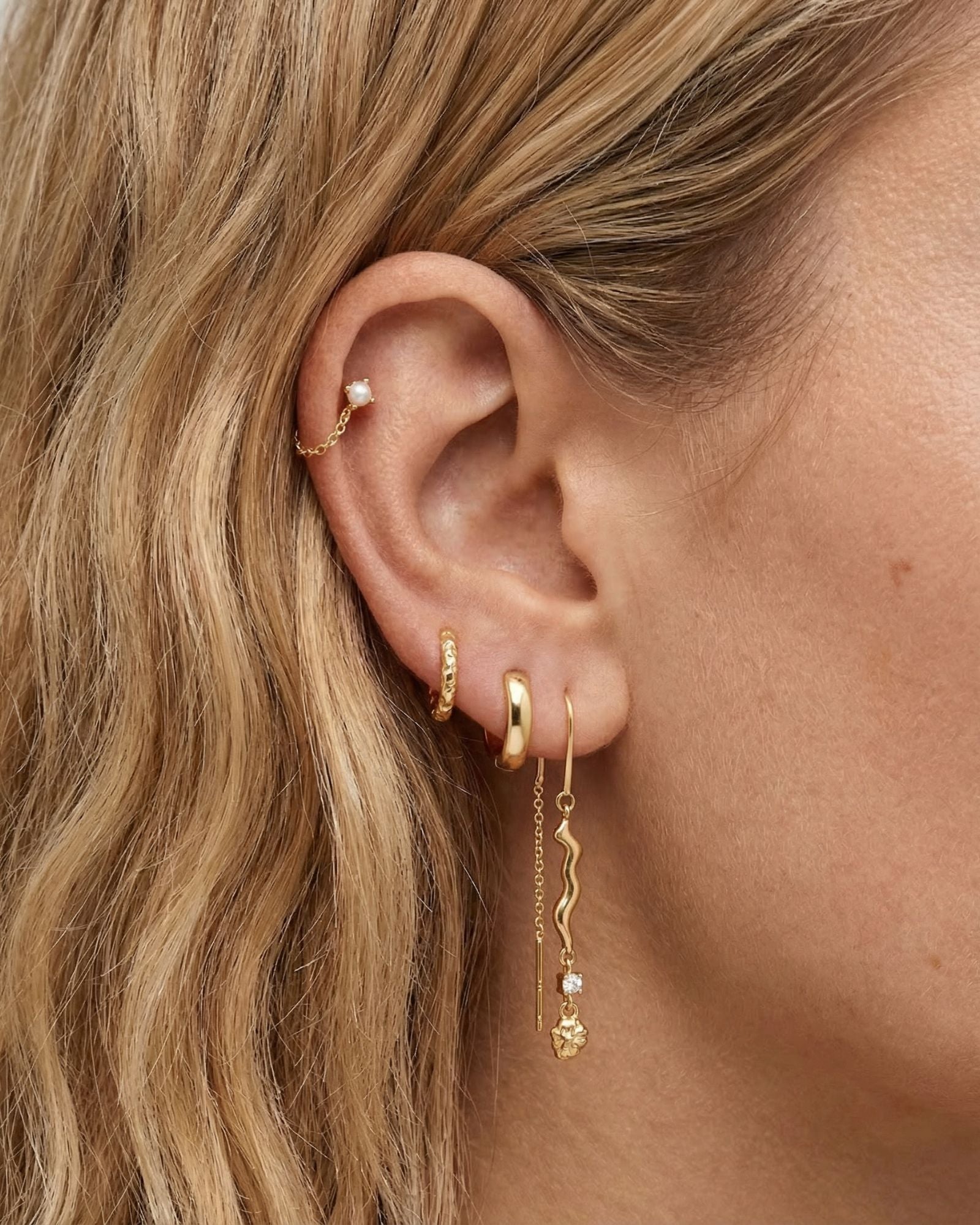 Close-up side profile of a blonde woman’s ear wearing layered gold jewellery, featuring a gold huggie hoop, a fine chain threader earring with a sculptural wavy bar and small crystal drop, and a delicate cartilage stud. Designed for women who like modern, minimal ear stacks, this image helps customers visualise how the gold drop earrings and threaders sit when styled together for everyday or elevated looks.