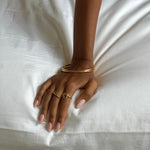 Woman’s hand resting on white fabric wearing multiple minimalist gold rings and slim gold bangles, featuring a twisted croissant-inspired gold stacking ring. The image highlights elegant everyday gold jewellery styled for women who want versatile, low-maintenance rings that layer easily and elevate simple outfits.