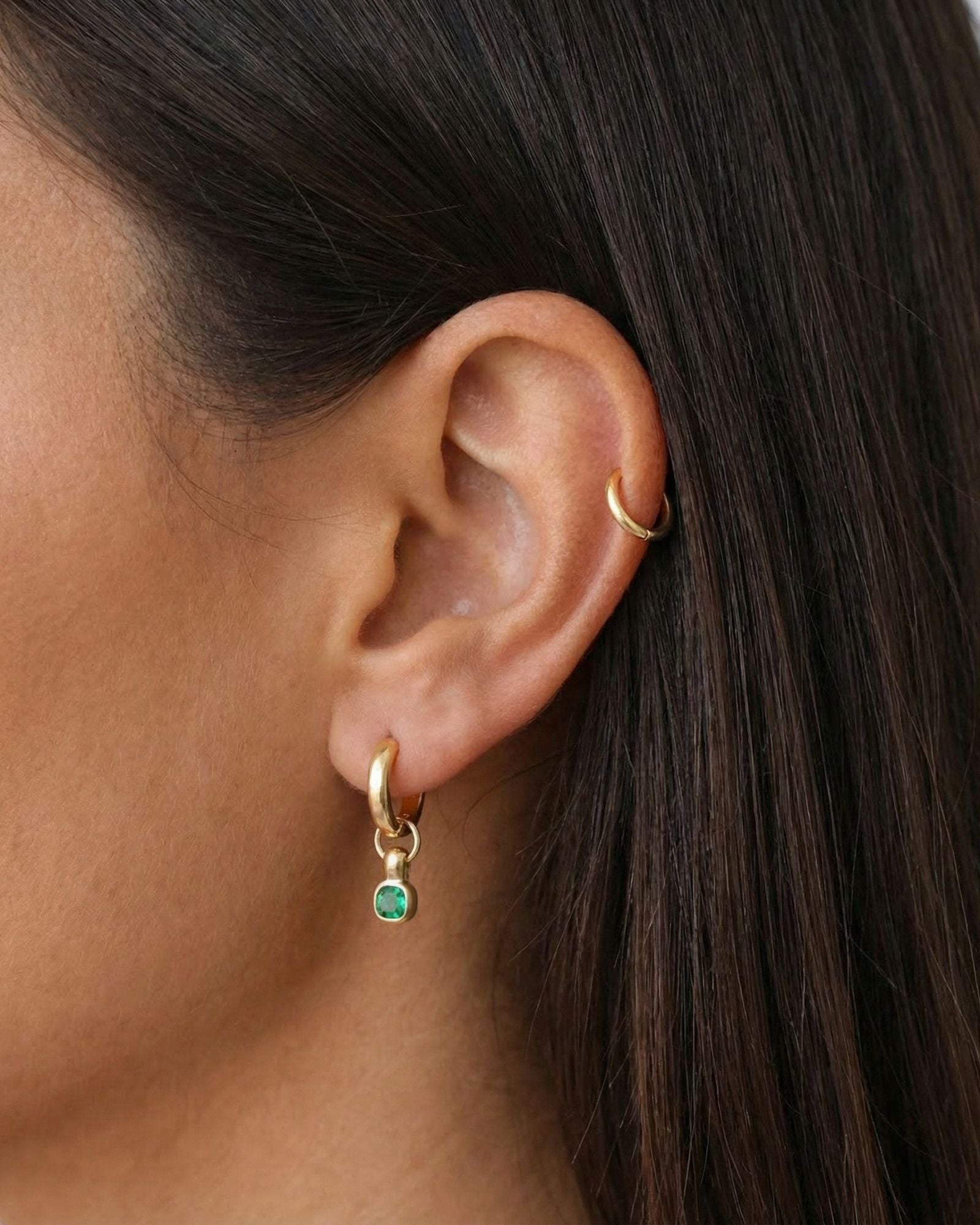 Close-up side profile of a woman’s ear wearing a gold huggie hoop earring with a small round green gemstone drop, paired with a minimal gold ear cuff worn higher on the cartilage. The image clearly shows the warm gold finish, the vibrant green stone detail, and how the drop earring moves naturally below the lobe. Designed for women who want subtle colour in their everyday jewellery, this image helps customers visualise how a gemstone huggie adds interest while remaining refined and easy to wear.
