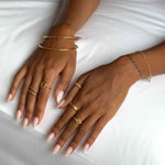 Close-up lifestyle image of a woman’s hands resting on white bedding, wearing stacked minimalist gold jewellery including slim gold bangles, delicate gold rings, and a fine gold chain bracelet. The image highlights a clean, elevated jewellery styling with polished gold finishes designed for women seeking timeless, everyday gold accessories that layer easily and add understated sophistication to daily outfits.