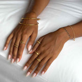 Close-up lifestyle image of a woman’s hands resting on white bedding, wearing stacked minimalist gold jewellery including slim gold bangles, delicate gold rings, and a fine gold chain bracelet. The image highlights a clean, elevated jewellery styling with polished gold finishes designed for women seeking timeless, everyday gold accessories that layer easily and add understated sophistication to daily outfits.