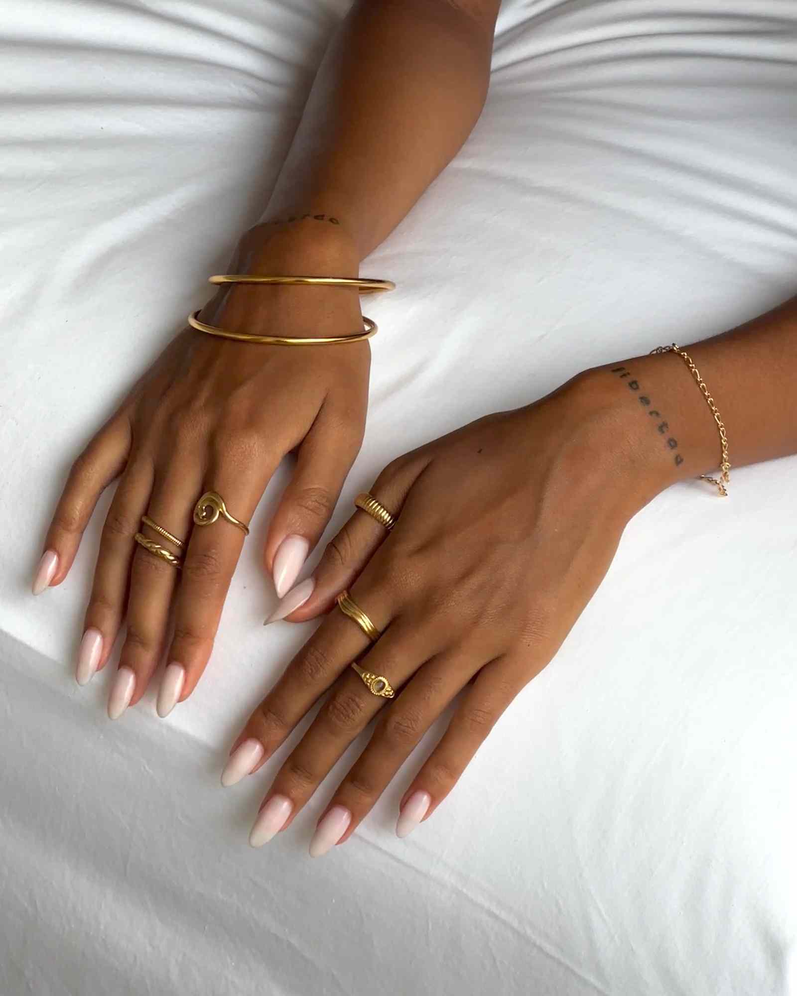 Close-up lifestyle image of a woman’s hands resting on white bedding, wearing stacked minimalist gold jewellery including slim gold bangles, delicate gold rings, and a fine gold chain bracelet. The image highlights a clean, elevated jewellery styling with polished gold finishes designed for women seeking timeless, everyday gold accessories that layer easily and add understated sophistication to daily outfits.