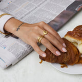 Product image showing a woman’s hand resting on a marble table beside a croissant and an open newspaper, wearing layered gold rings including a spiral gold ring, a wave-style stacking ring, and a slim gold band. Designed for women looking for everyday gold rings and minimalist stacking jewellery, this image highlights how versatile gold rings can elevate casual, lifestyle moments while offering effortless styling for daily wear.