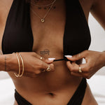 Model wearing layered gold jewellery including slim gold bangles, minimalist gold rings, and a delicate gold necklace, styled with a black bikini top. The image highlights everyday gold jewellery designed for women who want timeless, water-friendly pieces that elevate swimwear and warm-weather outfits while remaining lightweight and easy to wear.