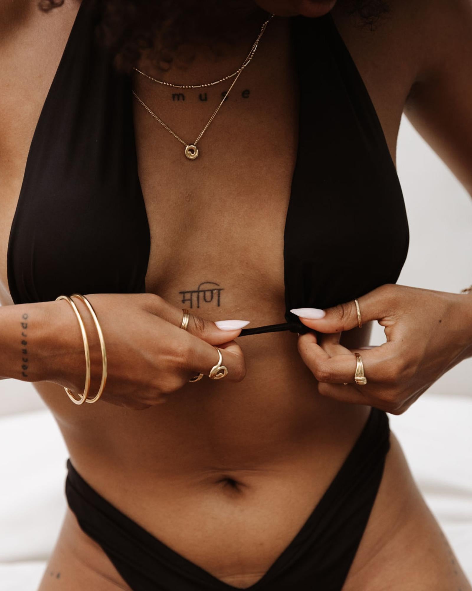 Model wearing layered gold jewellery including slim gold bangles, minimalist gold rings, and a delicate gold necklace, styled with a black bikini top. The image highlights everyday gold jewellery designed for women who want timeless, water-friendly pieces that elevate swimwear and warm-weather outfits while remaining lightweight and easy to wear.