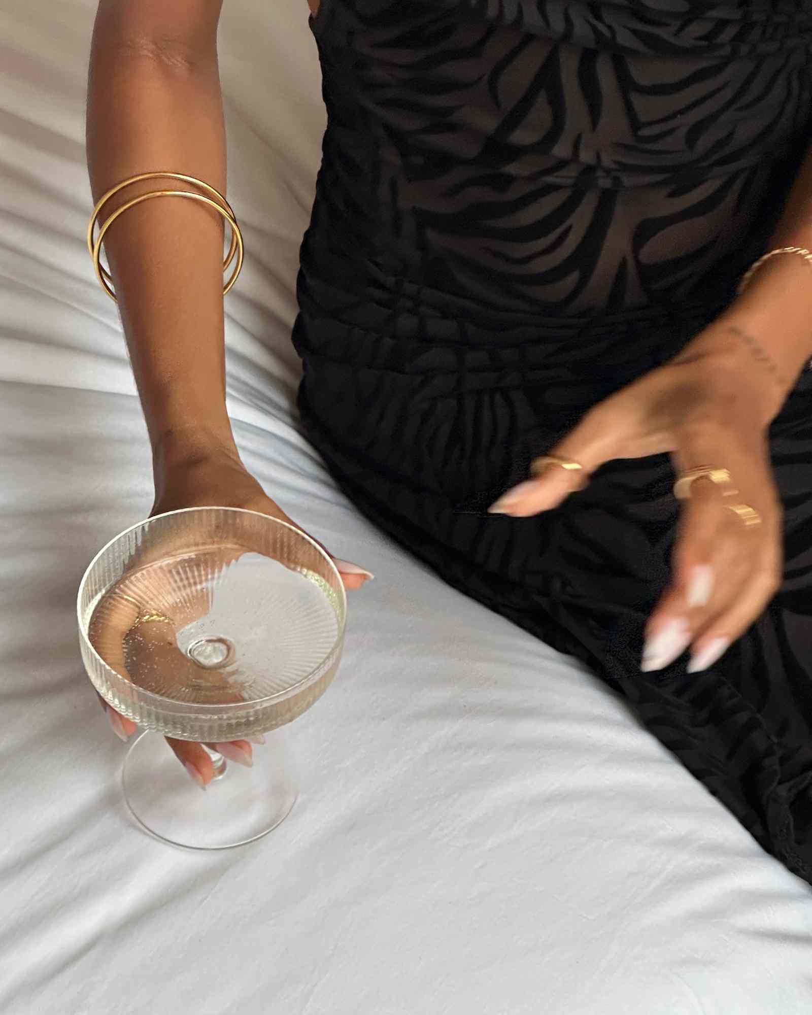 Woman wearing a slim minimalist gold bangle bracelet on her wrist while holding a clear glass coupe, styled with a black dress. The image shows a thin 14K gold filled bangle designed for everyday wear and stacking. Ideal for women seeking a lightweight, water-safe gold bracelet that adds subtle elegance to daily outfits and layered jewellery looks.