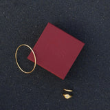 Flat lay product photo showing a slim gold bangle bracelet alongside two gold rings arranged on a dark textured surface, with a burgundy House of Slani jewellery box positioned nearby. The image highlights minimalist gold jewellery with clean lines and a polished finish, styled for women who prefer understated, everyday luxury. This photo helps shoppers visualise how a slim gold bangle and coordinating gold rings can be worn together or styled as a refined jewellery set for daily wear, stacking, or gifting.