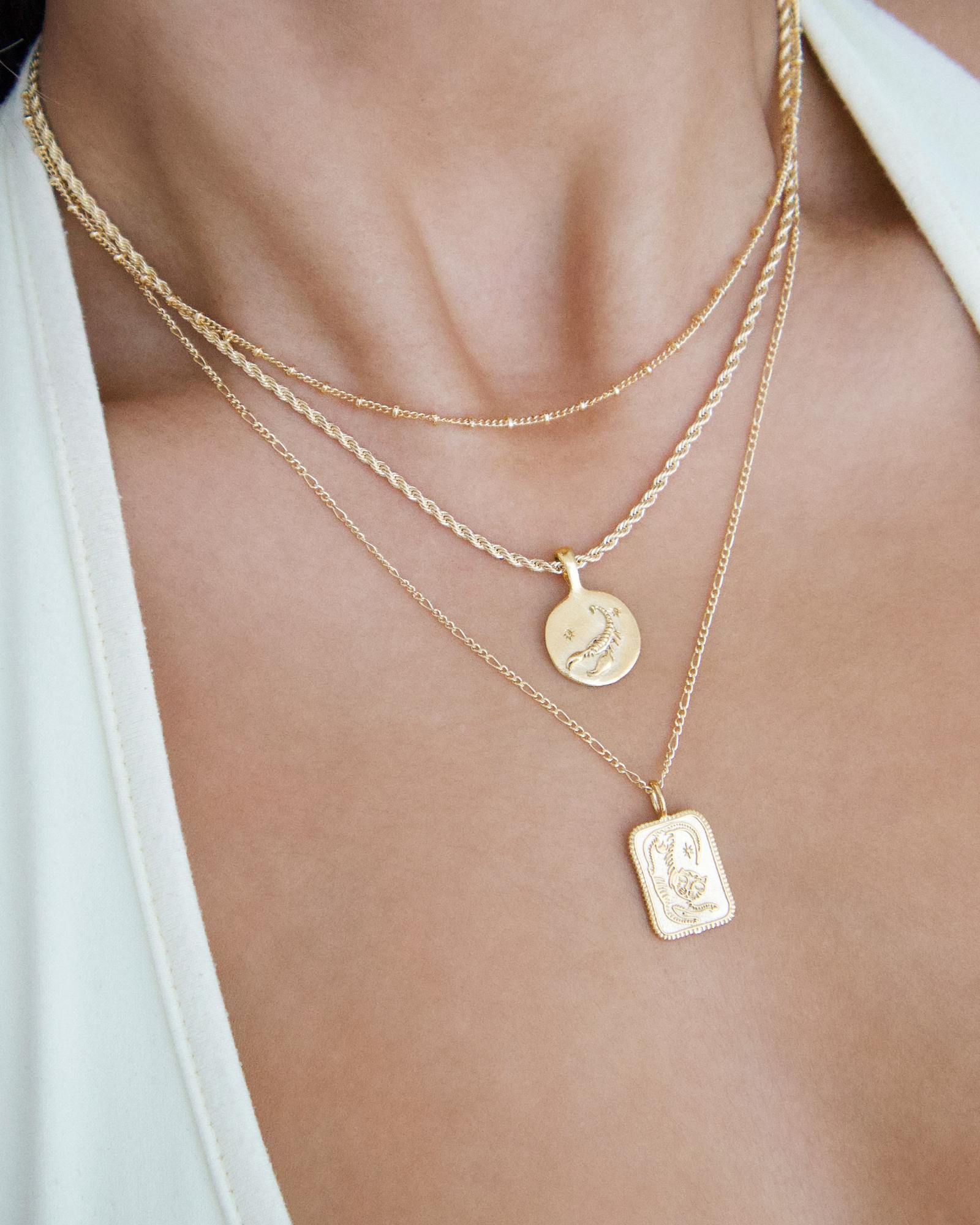 Layered gold necklaces worn on the neck and chest, featuring a fine gold rope chain paired with delicate gold chains and engraved pendant necklaces, including a round coin charm and a rectangular pendant. Styled against bare skin with a light neutral top, this image is for women seeking versatile gold layering necklaces that can be worn daily or dressed up. It helps show how lightweight gold chains create a balanced, elevated necklace stack without feeling heavy, making it easier to style everyday outfits 