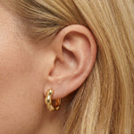 Close-up side profile of a woman’s ear wearing a sculptural gold huggie hoop earring with a softly rounded, organic shape and high-polish finish. The image clearly shows the earring’s fluid form, warm gold tone, and snug fit on the earlobe. Designed for women seeking a modern statement huggie that remains wearable for everyday use, this image helps customers understand scale, thickness, and how a bold yet refined gold hoop elevates a minimal look.