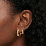 Close-up product photo of a woman’s ear with deep brown skin and natural textured hair, styled with layered gold huggie earrings. The lobe features a smooth, chunky gold huggie in the first piercing and a slimmer gold huggie set with small clear stones in the second piercing. This image is for customers looking for everyday gold earrings that balance bold shape with subtle sparkle. It helps show how pairing a statement gold huggie with a delicate stone-set hoop creates a polished, modern ear stack that feel