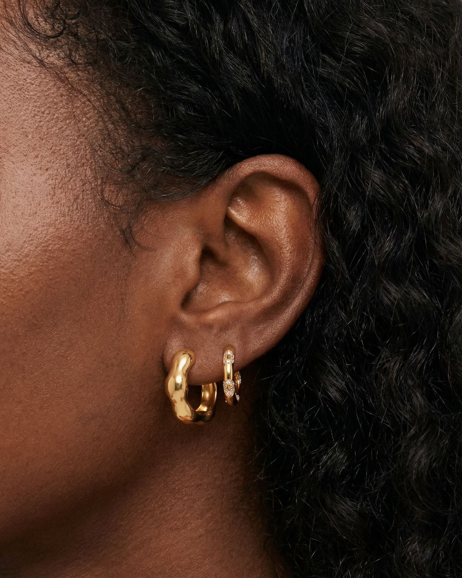 Close-up product photo of a woman’s ear with deep brown skin and natural textured hair, styled with layered gold huggie earrings. The lobe features a smooth, chunky gold huggie in the first piercing and a slimmer gold huggie set with small clear stones in the second piercing. This image is for customers looking for everyday gold earrings that balance bold shape with subtle sparkle. It helps show how pairing a statement gold huggie with a delicate stone-set hoop creates a polished, modern ear stack that feel