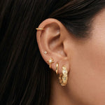 A person wearing a series of gold colored earrings, including medium sized hoop earrings with cubic zirconia, simple dainty flower stud earrings, and sun charm huggie hoops and a cartilage gold hoop.