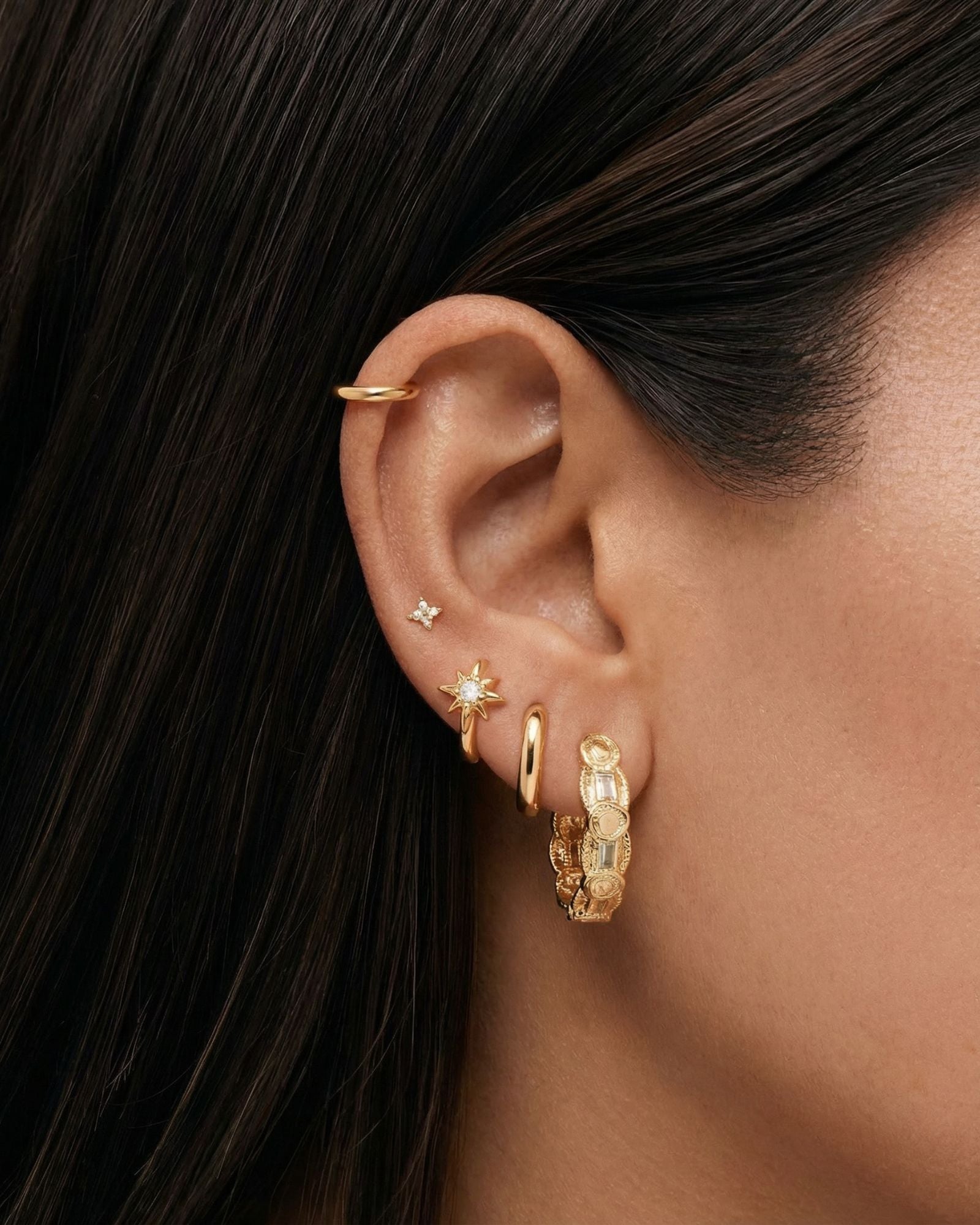 A person wearing a series of gold colored earrings, including medium sized hoop earrings with cubic zirconia, simple dainty flower stud earrings, and sun charm huggie hoops and a cartilage gold hoop.