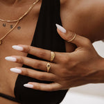 Lifestyle product photo showing a woman wearing layered gold rings on one hand, including a slim wave-style stacking ring, a delicate oval-detail gold ring, and a minimalist band, paired with fine gold necklaces and a black top. Designed for women seeking everyday gold jewellery and stacking ring inspiration, this image highlights how lightweight minimalist gold rings can be styled together for a refined, modern look that works for daily wear and elevated outfits.