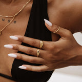 Lifestyle product photo showing a woman wearing layered gold rings on one hand, including a slim wave-style stacking ring, a delicate oval-detail gold ring, and a minimalist band, paired with fine gold necklaces and a black top. Designed for women seeking everyday gold jewellery and stacking ring inspiration, this image highlights how lightweight minimalist gold rings can be styled together for a refined, modern look that works for daily wear and elevated outfits.