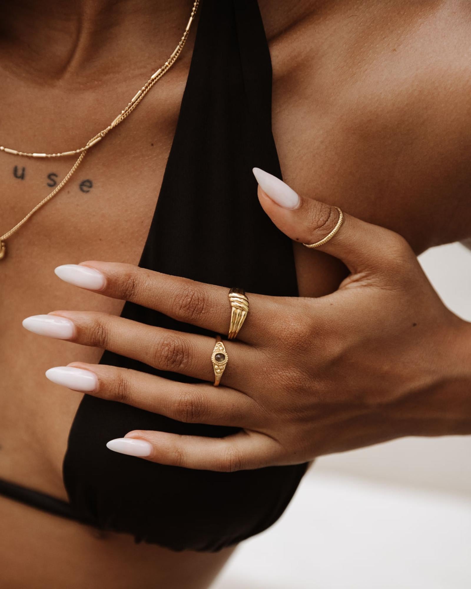 Lifestyle product photo showing a woman wearing layered gold rings on one hand, including a slim wave-style stacking ring, a delicate oval-detail gold ring, and a minimalist band, paired with fine gold necklaces and a black top. Designed for women seeking everyday gold jewellery and stacking ring inspiration, this image highlights how lightweight minimalist gold rings can be styled together for a refined, modern look that works for daily wear and elevated outfits.