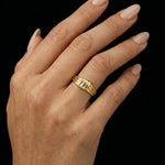 Close-up of a hand wearing a sculptural gold ring with a layered, organic band design, shown against a black background. The ring sits on the finger with a polished finish and softly contoured shape, highlighting its statement yet refined aesthetic. This image is for customers seeking an elevated essential ring that adds visual interest without overpowering an outfit, suitable for everyday wear, styling confidence, and modern personal expression.
