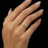 Close-up of a hand wearing a sculptural gold ring with a layered, organic band design, shown against a black background. The ring sits on the finger with a polished finish and softly contoured shape, highlighting its statement yet refined aesthetic. This image is for customers seeking an elevated essential ring that adds visual interest without overpowering an outfit, suitable for everyday wear, styling confidence, and modern personal expression.