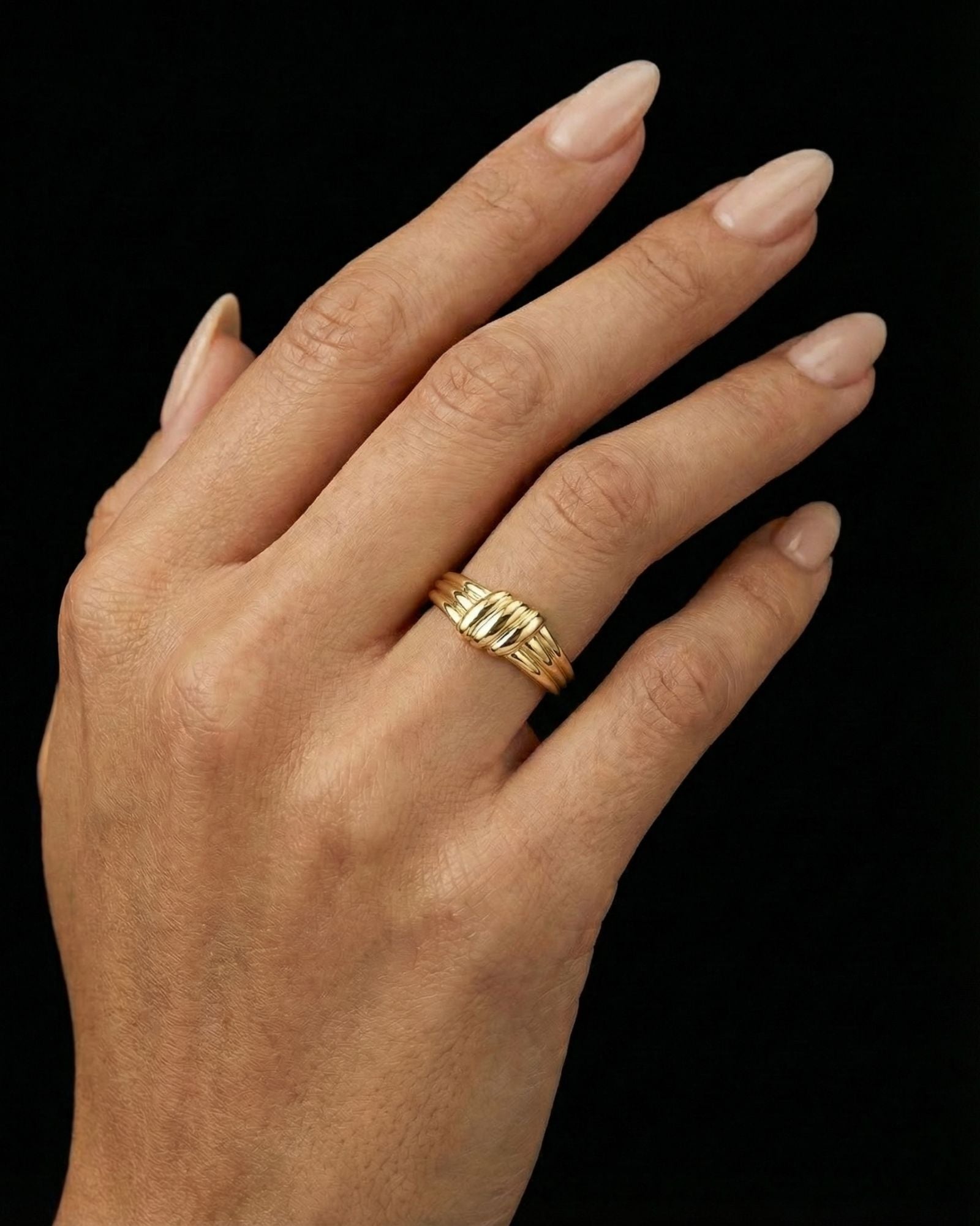 Close-up of a hand wearing a sculptural gold ring with a layered, organic band design, shown against a black background. The ring sits on the finger with a polished finish and softly contoured shape, highlighting its statement yet refined aesthetic. This image is for customers seeking an elevated essential ring that adds visual interest without overpowering an outfit, suitable for everyday wear, styling confidence, and modern personal expression.