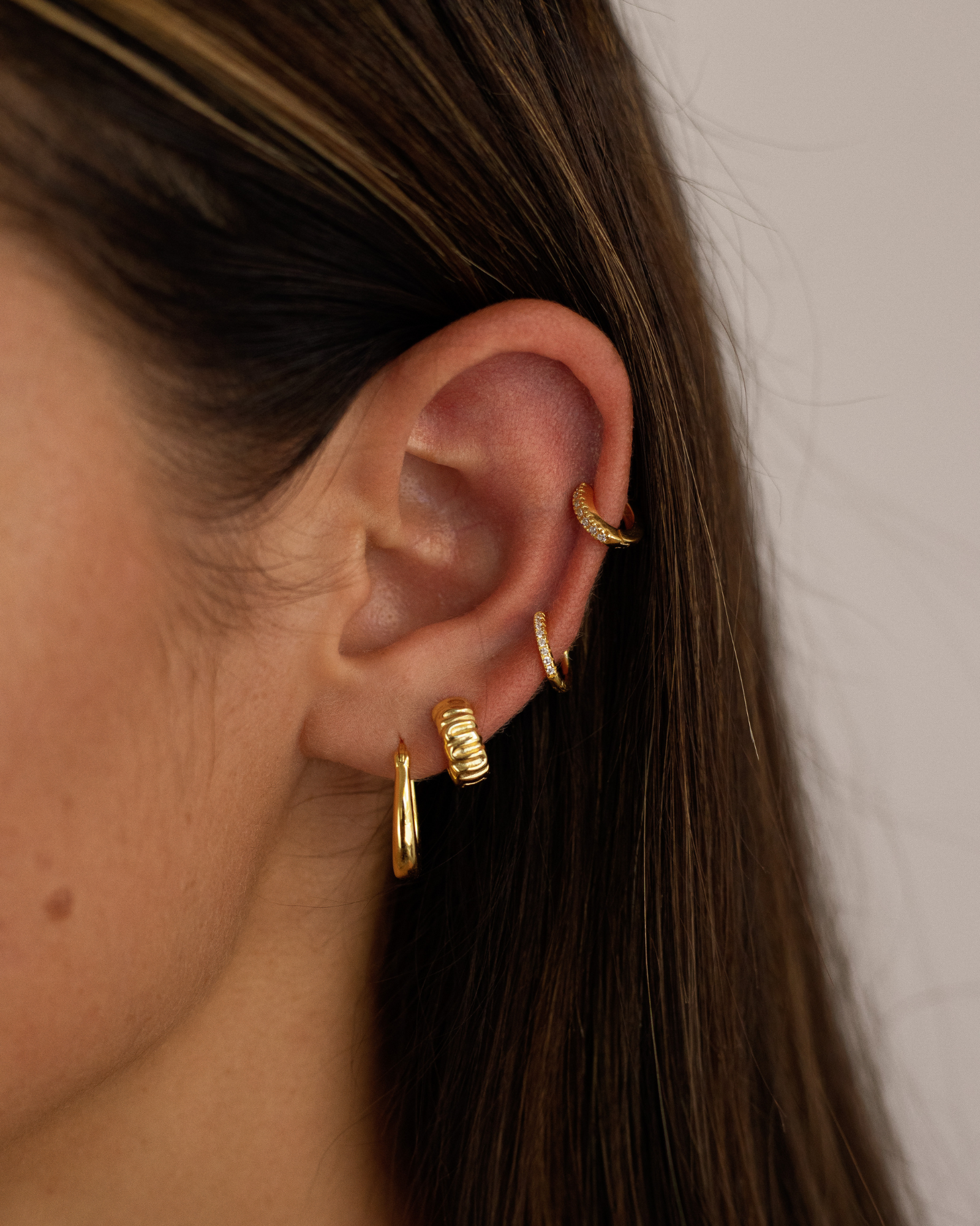 Close-up side view of a woman’s ear styled with layered gold earrings, including small gold huggie hoops, a textured gold hoop in the lobe, and slim pavé huggie hoops in the helix and upper lobe. Designed for women looking for stackable gold earrings, helix and lobe jewelry, and everyday pieces that solve the challenge of creating a balanced, cohesive ear stack with multiple piercings while remaining refined and wearable for daily use.