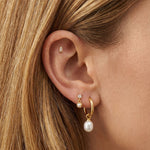 Close-up of an ear styled with minimalist gold jewellery, featuring a small rectangular gold stud in the cartilage, a delicate crystal and pearl drop stud, and a gold hoop earring with a freshwater pearl charm. Designed for women seeking elegant gold earrings for everyday wear, this image shows how pearl drop earrings, gold hoops, and subtle stud earrings can be layered for a refined ear stack that suits sensitive ears, first or second piercings, and a polished, modern jewellery look.