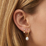 Close-up of an ear styled with minimalist gold jewellery, featuring a small rectangular gold stud in the cartilage, a delicate crystal and pearl drop stud, and a gold hoop earring with a freshwater pearl charm. Designed for women seeking elegant gold earrings for everyday wear, this image shows how pearl drop earrings, gold hoops, and subtle stud earrings can be layered for a refined ear stack that suits sensitive ears, first or second piercings, and a polished, modern jewellery look.