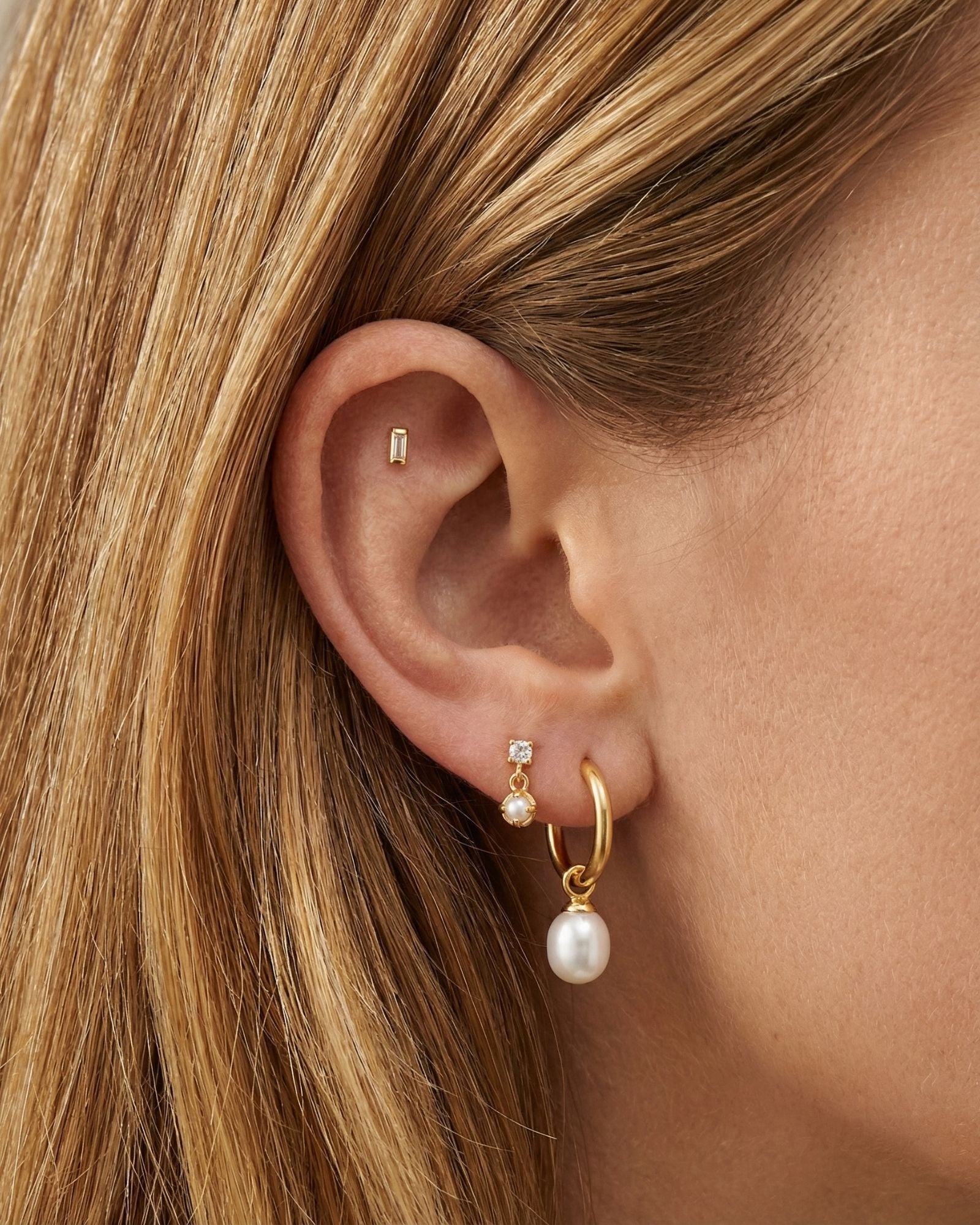 Close-up of an ear styled with minimalist gold jewellery, featuring a small rectangular gold stud in the cartilage, a delicate crystal and pearl drop stud, and a gold hoop earring with a freshwater pearl charm. Designed for women seeking elegant gold earrings for everyday wear, this image shows how pearl drop earrings, gold hoops, and subtle stud earrings can be layered for a refined ear stack that suits sensitive ears, first or second piercings, and a polished, modern jewellery look.