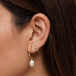 Close-up lifestyle image of a woman’s ear styled with layered gold jewellery, featuring a small gold crystal stud in the upper lobe, a delicate gold huggie with clear stones, and a gold hoop earring with a dangling freshwater pearl charm. Designed for women who enjoy refined ear stacking and modern pearl jewellery, this image shows how gold pearl drop earrings and minimal studs can be layered for an elegant yet wearable everyday look, ideal for adding softness and sophistication to casual, work, or occasion