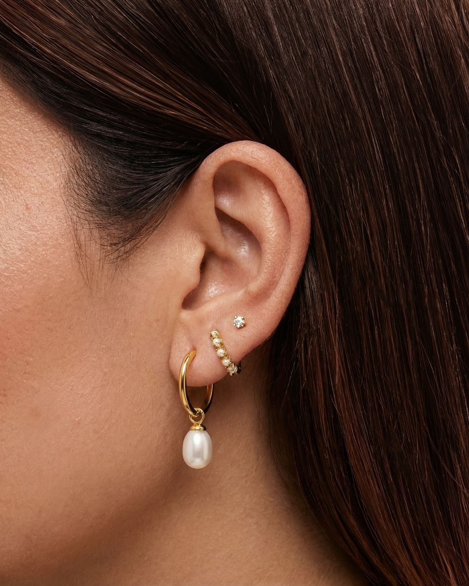 Close-up lifestyle image of a woman’s ear styled with layered gold jewellery, featuring a small gold crystal stud in the upper lobe, a delicate gold huggie with clear stones, and a gold hoop earring with a dangling freshwater pearl charm. Designed for women who enjoy refined ear stacking and modern pearl jewellery, this image shows how gold pearl drop earrings and minimal studs can be layered for an elegant yet wearable everyday look, ideal for adding softness and sophistication to casual, work, or occasion
