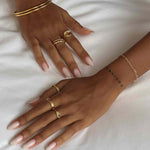 Product image showing two hands wearing layered gold rings, including a minimalist gold wave stacking ring, a small oval signet ring, and slim gold bands, paired with gold bangles and a fine chain bracelet on a white fabric background. Designed for those searching for gold stacking rings, minimalist gold jewellery, and everyday gold accessories, this image highlights coordinated gold pieces that solve the need for effortless layering, cohesive styling, and refined daily wear.