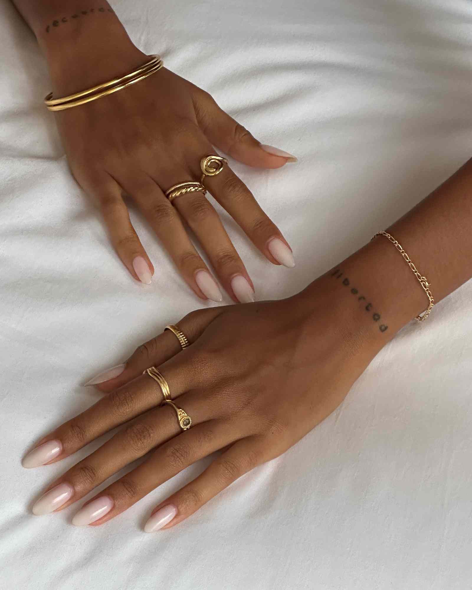 Product image showing two hands wearing layered gold rings, including a minimalist gold wave stacking ring, a small oval signet ring, and slim gold bands, paired with gold bangles and a fine chain bracelet on a white fabric background. Designed for those searching for gold stacking rings, minimalist gold jewellery, and everyday gold accessories, this image highlights coordinated gold pieces that solve the need for effortless layering, cohesive styling, and refined daily wear.
