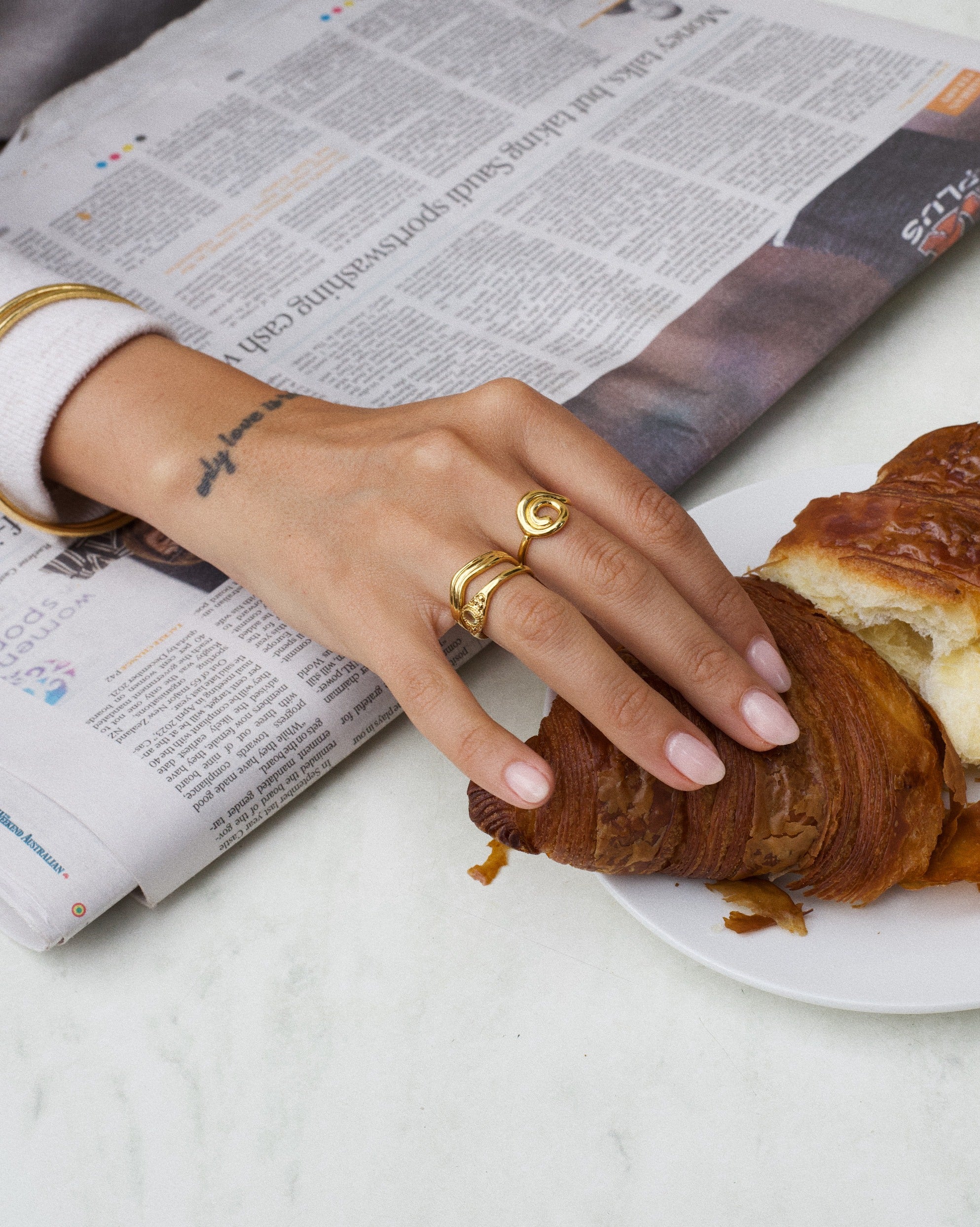 Product image of a woman’s hand wearing stacked gold rings, including an organic spiral gold ring and a slim textured band, resting beside a croissant on a plate and a newspaper. Designed for women seeking everyday gold jewellery, this image shows how minimalist gold rings can be styled effortlessly for daily routines, offering a versatile and understated option for casual, refined wear.