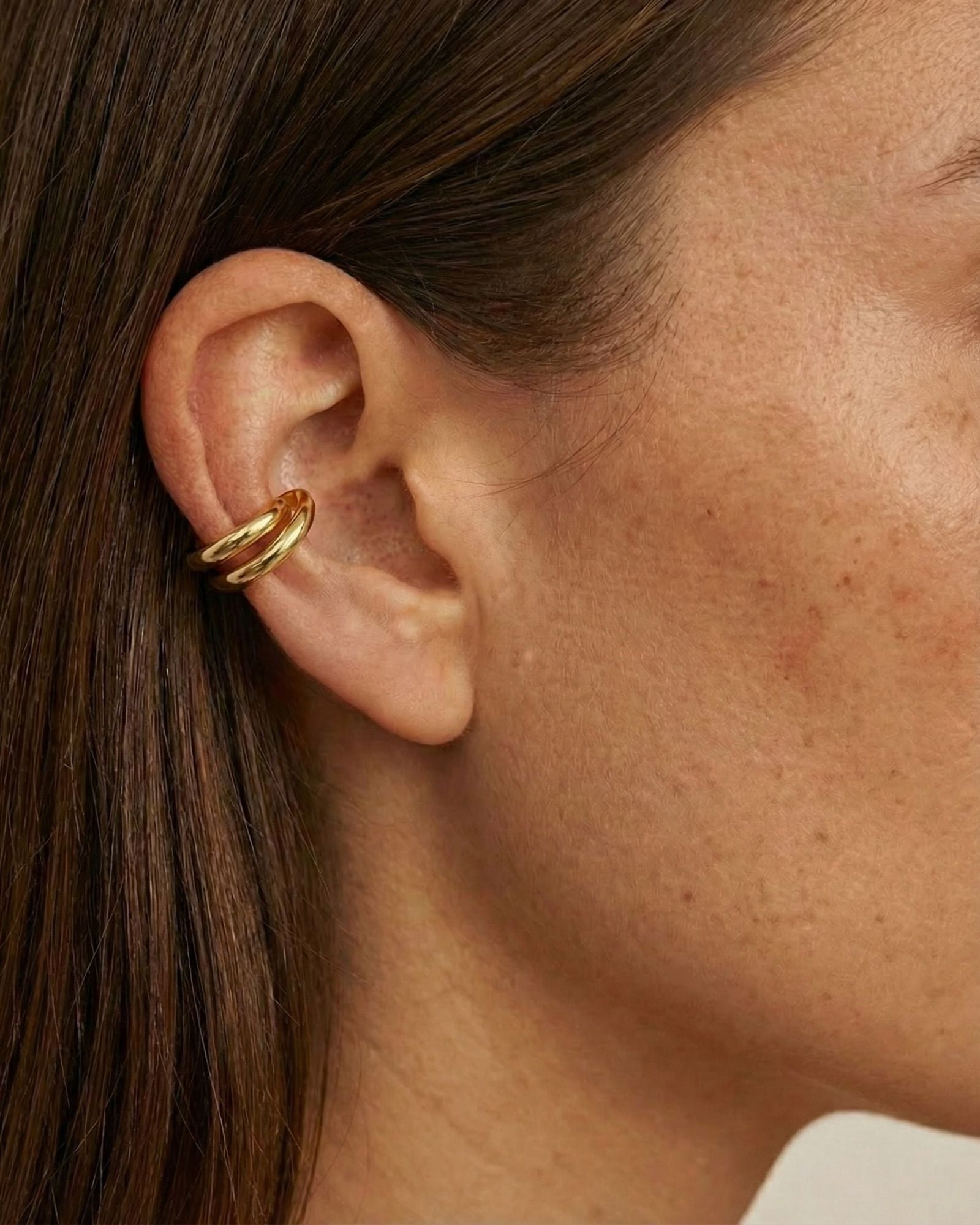 Close-up side profile of a woman wearing a polished gold ear cuff with a smooth, double-band design, positioned on the outer ear cartilage without a piercing. The image clearly shows the cuff’s rounded shape, warm gold tone, and snug fit against natural skin and brown hair. Designed for women who want an easy, non-pierced ear accessory, this image helps customers understand how a minimalist gold ear cuff sits on the ear and adds subtle definition to an everyday look.