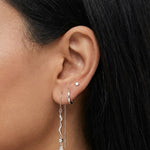 Close-up side profile of a dark-haired woman’s ear styled with silver jewellery, including a silver hoop, a slim chain threader earring with a wavy bar and crystal detail, and a small stud piercing. Intended for women seeking clean, contemporary silver earrings, this image shows scale, length, and layering to help customers understand how the silver drop earrings move and wear on the ear.