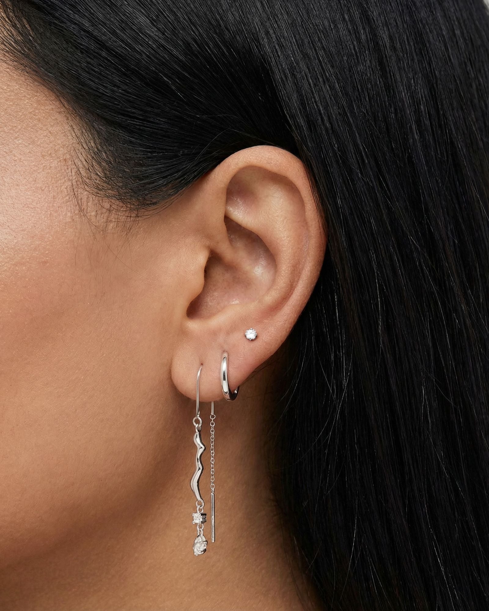 Close-up side profile of a dark-haired woman’s ear styled with silver jewellery, including a silver hoop, a slim chain threader earring with a wavy bar and crystal detail, and a small stud piercing. Intended for women seeking clean, contemporary silver earrings, this image shows scale, length, and layering to help customers understand how the silver drop earrings move and wear on the ear.
